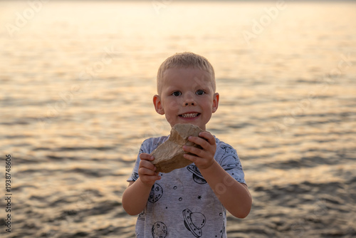 Cheerful blond boy smiling broadly at camera while proudly showing found rock stone standing on sandy beach with warm glowing sea at sunset. Childhood pride, outdoor discovery, happy seaside memories.