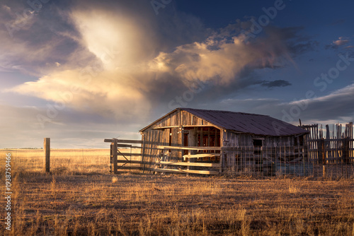 Tablou pe pânză An old, abandoned farm with a wooden fence under a stormy sky at sunset in the rural countryside