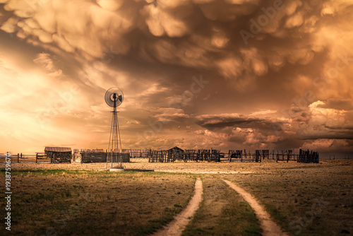 An old, abandoned farm and a spinning windmill under a stormy sky at sunset in the rural countryside.