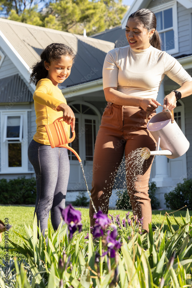 Fototapeta premium Mother and school-age daughter watering purple iris in yard using orange and white watering cans