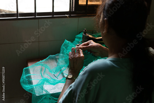 Bilde på lerret Back view of an elderly woman meticulously sewing a green fishing net by the win