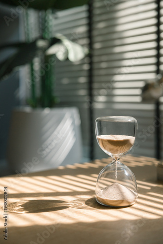 Sand flows through the glass beads in the hourglass, casting beautiful shadows on the wooden table, while natural light from the window signifies the passage of time.