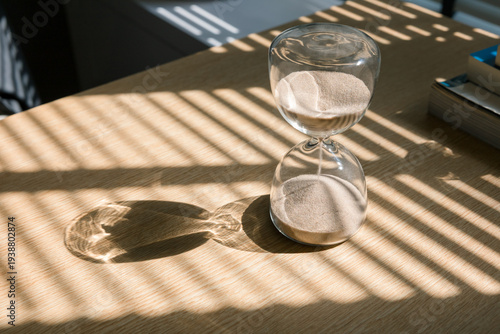 Sand flows through the glass spheres of an hourglass on a wooden desk, casting beautiful shadows in the natural light from the window, measuring the passage of time the concept of timing. 