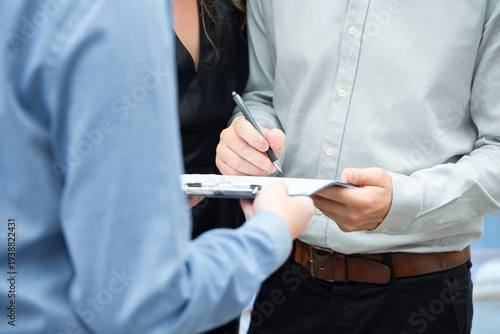 Close up business transaction showing client signing car purchase documents on clipboard while dealership consultant holds paperwork beside new vehicle in showroom environment.
