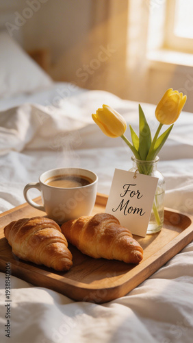 Breakfast in bed with croissants coffee and yellow tulips. Mother's Day tray with food and For Mom card in sunny bedroom