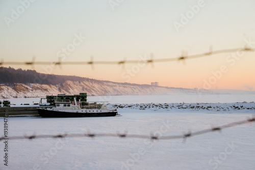 Boat moored for winter behind barbed wire on a frozen river at sunset