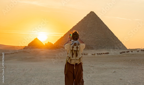Fotografie Tourist takes a photo of the pyramids in Egypt during sunset while camels walk i