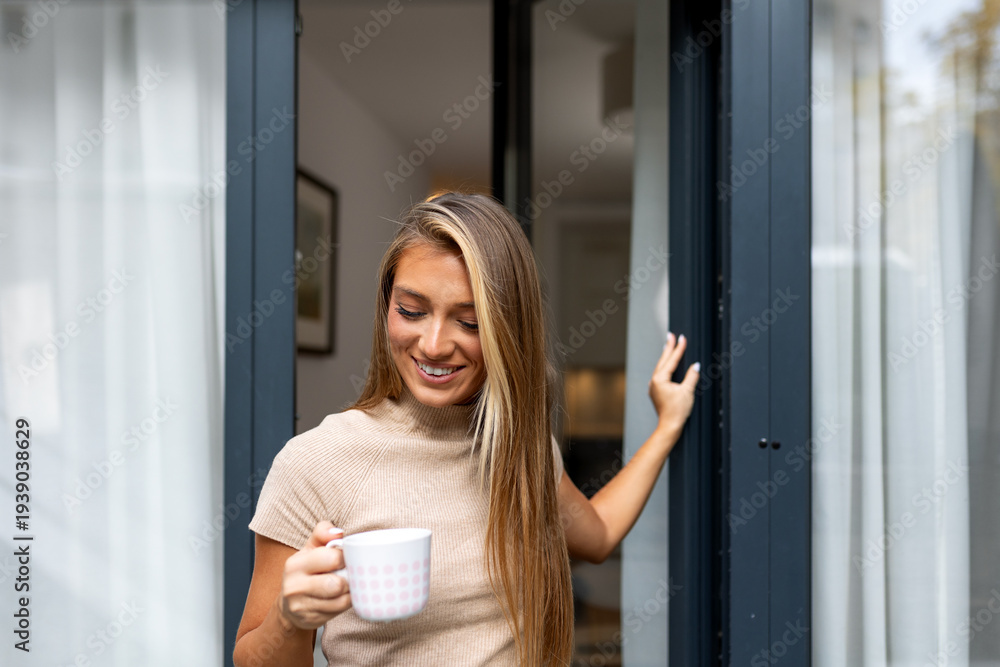 Fototapeta premium Smiling woman standing by balcony door with coffee cup. Morning relaxation, home comfort, and lifestyle concept. Happy female holding mug and looking down. Peaceful moment in modern apartment home.