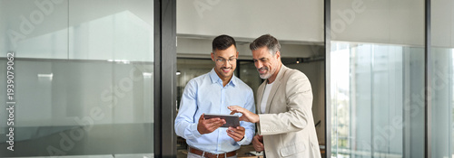 Business team of two happy successful professional business men executive working together standing in office using digital tablet computer technology managing data standing in corporate office.