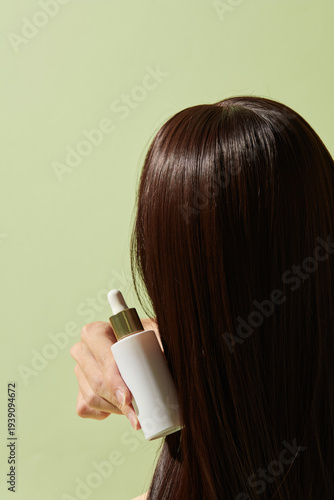 Woman holding blank white haircare bottle near long glossy hair on pastel green background, product mockup for shampoo or conditioner branding