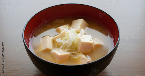 A close up shot displays a dark red bowl filled with a clear broth containing cubed tofu and thinly sliced scallions high quality professional detailed modern