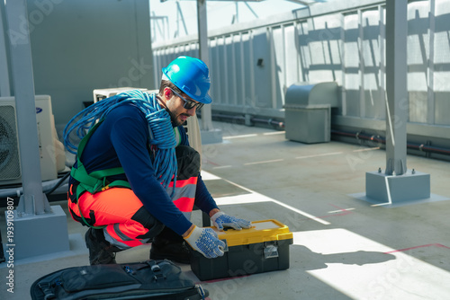Bilde på lerret Professional HVAC technician wearing protective helmet and safety harness crouches on commercial building rooftop opening toolbox near air conditioner condenser units preparing repair equipment