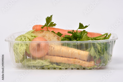 A variety of fresh soup vegetables including carrots, parsley, onion and parsnip packed in a transparent plastic container on a white background