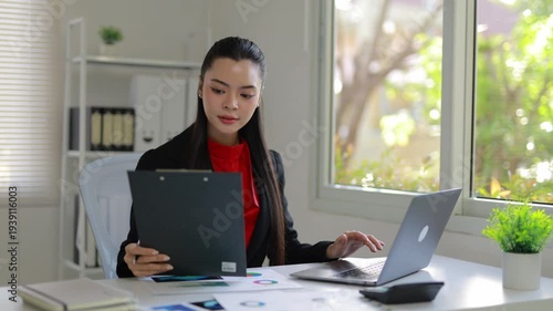 Professional Asian woman working in a modern office, reviewing financial documents and using a laptop to analyze company performance data