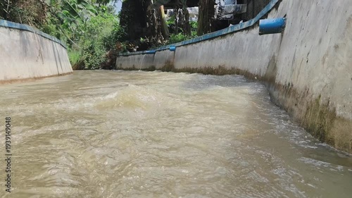 A flow of murky water through a concrete channel lined with blue trim, surrounded by lush green vegetation.