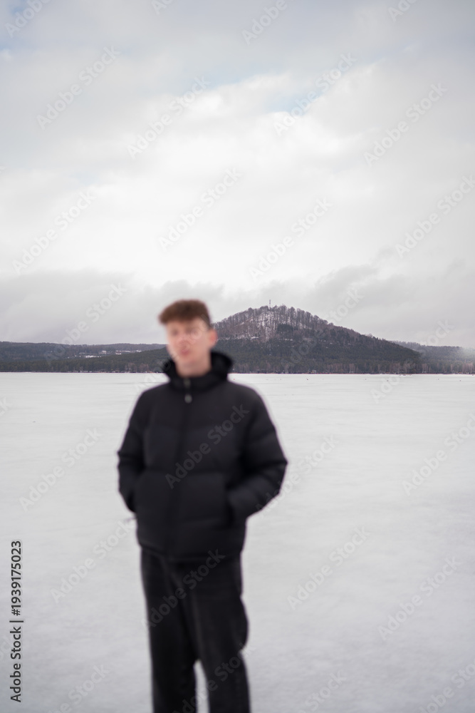 Fototapeta premium View of snowy hill across Macha Lake with blurred man in foreground. Winter landscape in Czech Republic. Man standing near frozen Macha Lake in Czech Republic looking at distant hill. Winter travel an