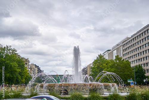 Photos A scenic urban view featuring a large ornate fountain as the central focal point, with multiple arcing water jets creating elegant sprays and a tall central column of water shooting skyward