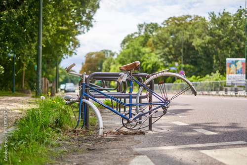 Old damaged blue bicycle abandoned and locked to a metal bike rack on a Paris street, missing front wheel, bent rear wheel with rusted spokes