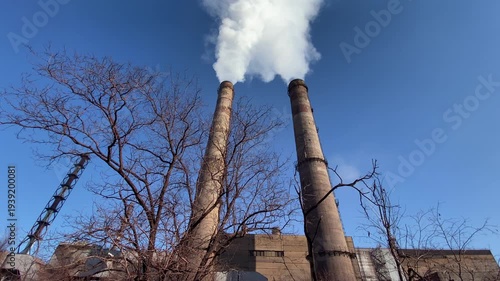 Two factory smokestacks releasing white smoke above industrial building and bare trees. Concept of air pollution, industry emissions and environmental impact.