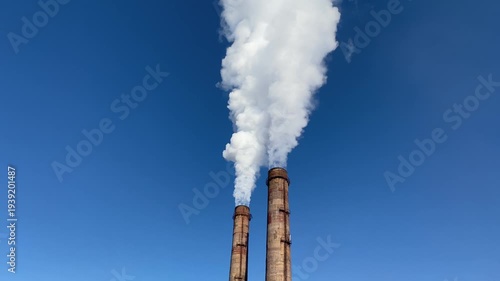 Two factory smokestacks releasing thick white smoke into clear blue sky. Industrial emissions, air pollution and environmental impact concept.