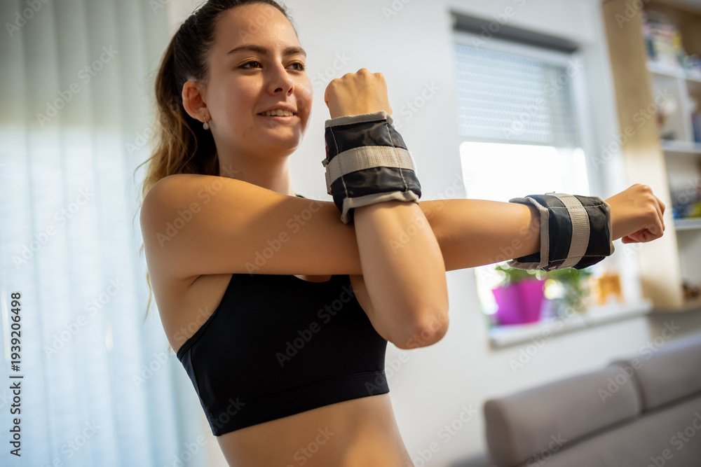 Fototapeta premium Young woman in sportswear stretching her shoulder with wrist weights at home.