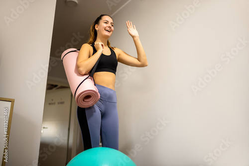 Smiling young woman in sportswear carrying a yoga mat and waving while entering a gym or pilates studio.