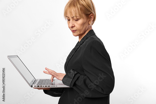 Mature woman in black blazer working on laptop, focused on screen with neutral expression. Isolated on white background, concept for business, technology, and professionalism.