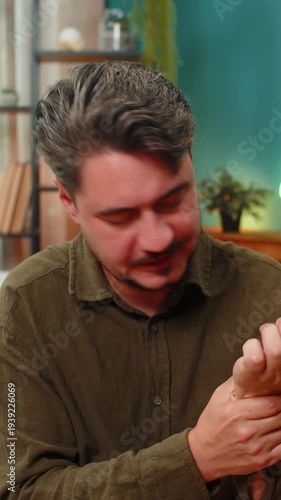 Middle-aged man at home massages aching wrist feels sharp strain injury seeks relief comfort. Young adult guy on sofa rubs hand muscles tense worries about joint health repetitive stress causing pain