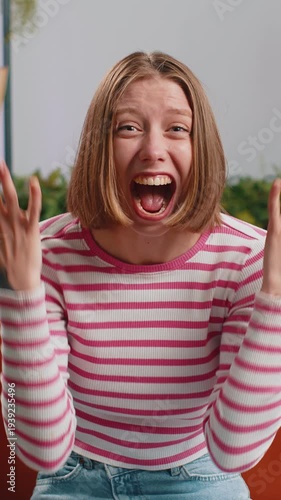 Happy young brunette woman shouting, celebrating success, winning, goal achievement good news raising fists in gesture I did it, lottery luck at home apartment. Lady in living room sitting on couch