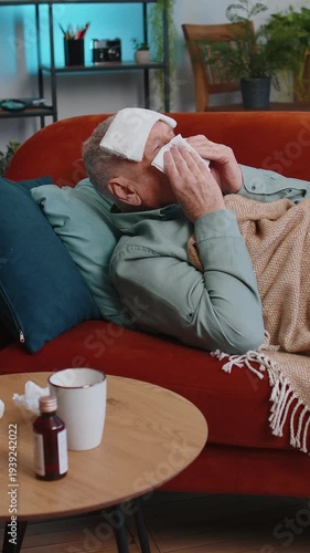 Grandfather ill man suffering from cold or allergy pain lying on couch in living room at home. Sick elderly guy with towel on head blows sneezes wipes snot into napkin. Coronavirus quarantine pandemic