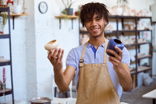 Smiling adult man holding pottery vases in studio during daytime