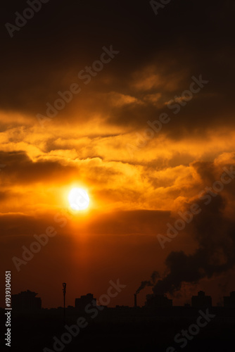 Dramatic orange sunset. Vibrant colors of the evening orange glow in the cloudy sky over a modern city with smoking pipes. A spectacular atmospheric phenomenon. Ecology. vertical stock photo 2x3