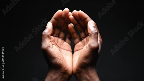 A close-up shows two dark-skinned hands, gently cupped and open against a stark black background. This profound, expressive gesture conveys universal themes of offering, receiving, prayer, hope, and c