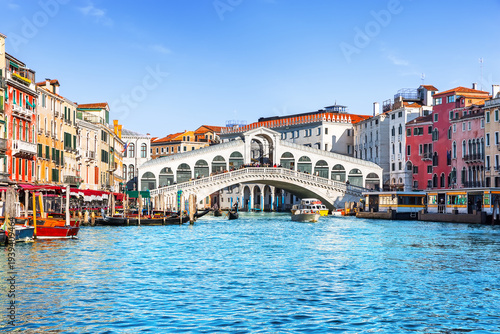 Famous Rialto Bridge spanning the Grand Canal in Venice, Italy, surrounded by historic colorful buildings and turquoise water under clear blue sky. 