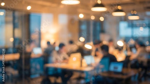 Softly blurred lights create a captivating bokeh effect in this modern office interior, where indistinct figures are diligently working at their desks, evoking a sense of focused activity.
