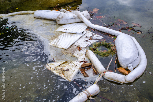 Oil containment booms are used to manage water pollution in a lake during winter near a small town in the background with mountains visible
