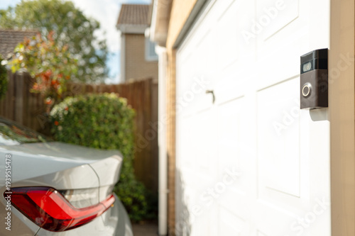 Shallow focus of a popular, battery powered wireless doorbell and integral camera seen located on a driveway, near a side gate entrance. A parked car is on the driveway.