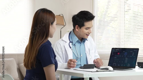 Asian male doctor consulting with female patient while using calculator and laptop in bright clinic