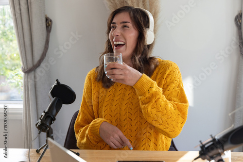 Female host laughing holding glass in yellow sweater headphones mic recording on laptop in studio