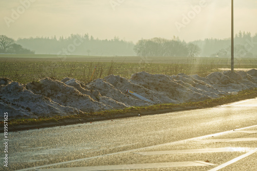 Sunny road with dirty sand snow banks beside green field in warm light