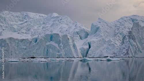 A vast glacier and icebergs reflected in calm water under a cloudy, overcast sky, depicting a serene polar landscape.