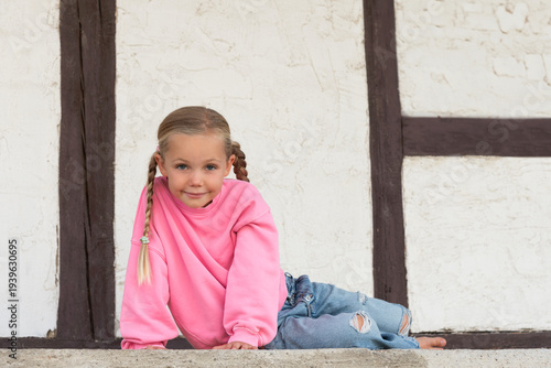 6 year old girl with pink jumper and jeans sitting sideways in front of old white wall with wooden beams looking curious
