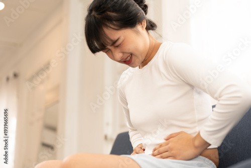 Foto Asian woman sitting on sofa holding stomach with painful expression, suffering from abdominal pain at home, concept of stomachache, illness, health problem