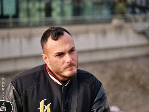 Portrait of a middle-aged man with a beard wearing a black varsity jacket sitting outdoors. Contemplative male face close-up, urban lifestyle, thoughtful expression, casual streetwear style, 