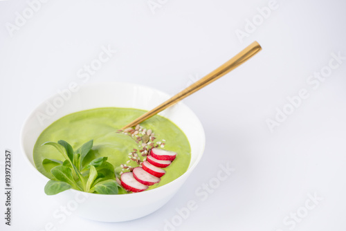 Healthy green spring soup in a white bowl garnished with sliced radishes and sprouts, accompanied by a golden spoon against a clean white background