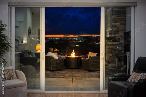 Bild auf Leinwand A young woman relaxes by a flaming fire pit on a garden patio overlooking the city lights of Spokane, Washington at night