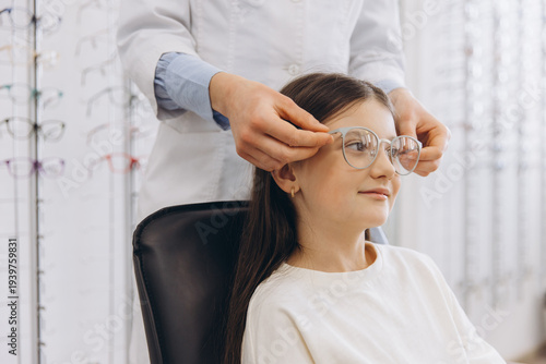 Optometrist helping young girl trying on new glasses for vision correction at optical shop