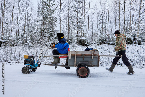 Two wheel tractor with trailer travels along forest road driven by farmer second farmer walks behind in snowy winter woodland.