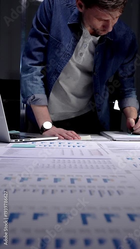 Entrepreneur standing at a desk in a dark office, studying financial reports and charts by lamp light while working on a laptop, focused on strategic planning and data analysis