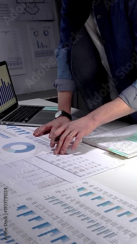 Person analyzing financial data, reviewing business reports and charts on a desk during a late work session, using a laptop for statistics and market research